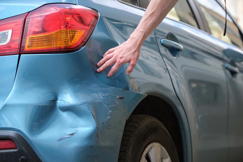 Person examining a dent and scratches on the rear side of a blue car after a minor collision.