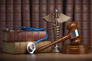 A gavel, a stack of medical books, and a stethoscope sit on a desk infant of a bookshelf.