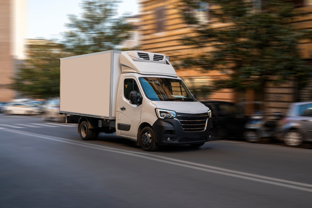 White delivery truck driving through a city street surrounded by cars and buildings.