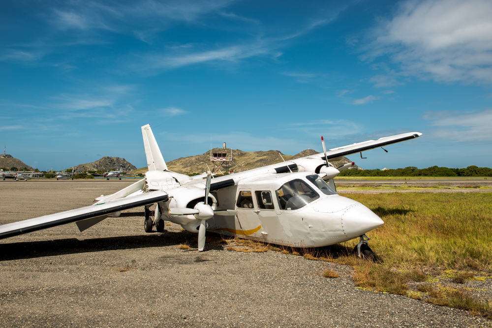Small white airplane with visible damage resting on the ground beside a runway under clear blue skies.