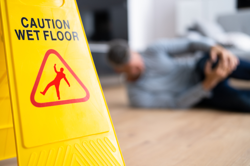 “Caution Wet Floor” sign with injured man in the background, illustrating slip and fall accident hazards.