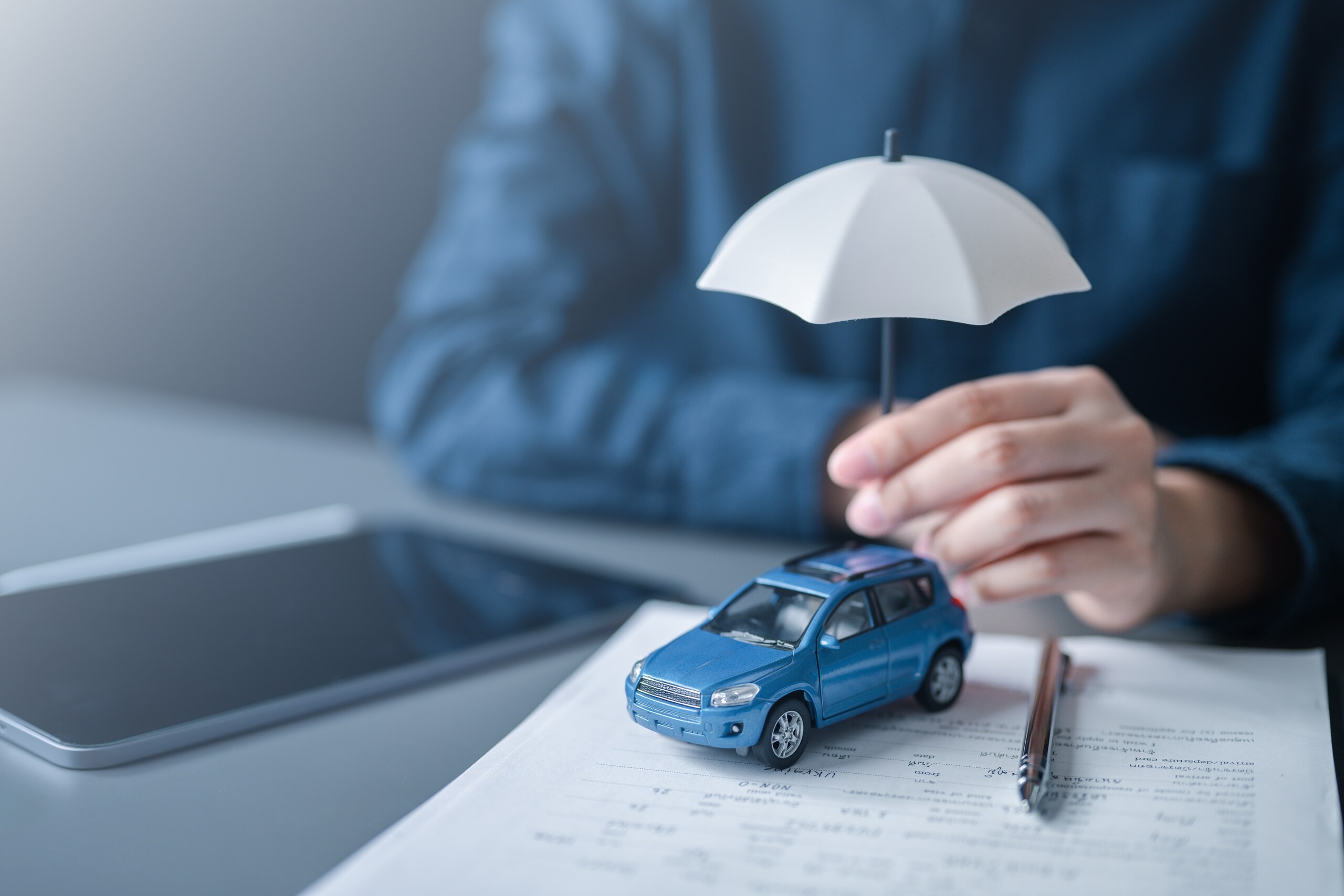 A person holding a small umbrella over a model car on insurance paperwork, illustrating liability coverage in Texas.