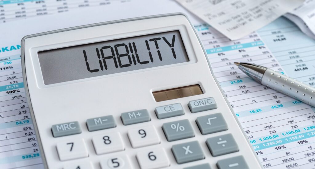 A close-up of a calculator displaying the word "LIABILITY" on a desk with financial documents and a pen.