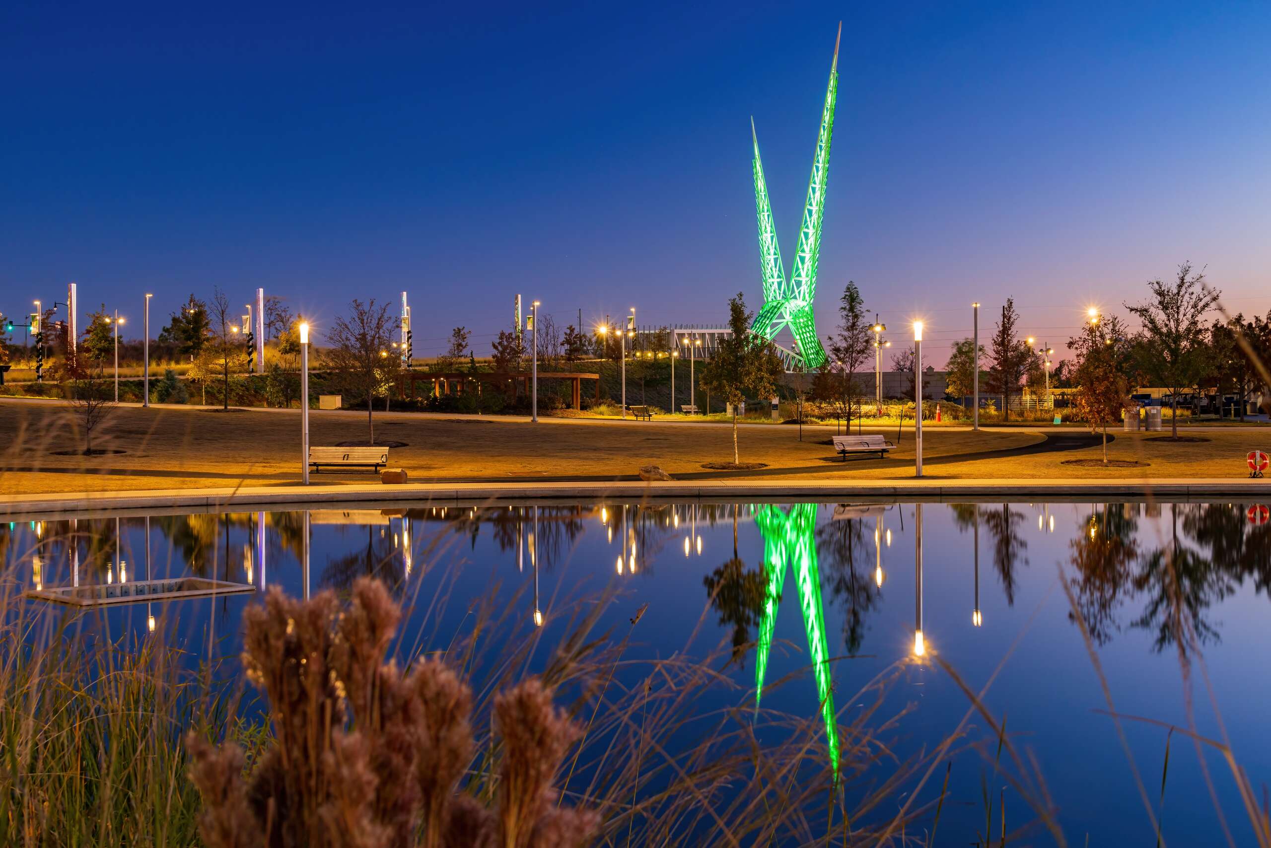 The illuminated green Skydance Bridge at dusk, with its reflection visible in a park pond, providing local geographical context for Oklahoma City Truck Accident Attorneys.
