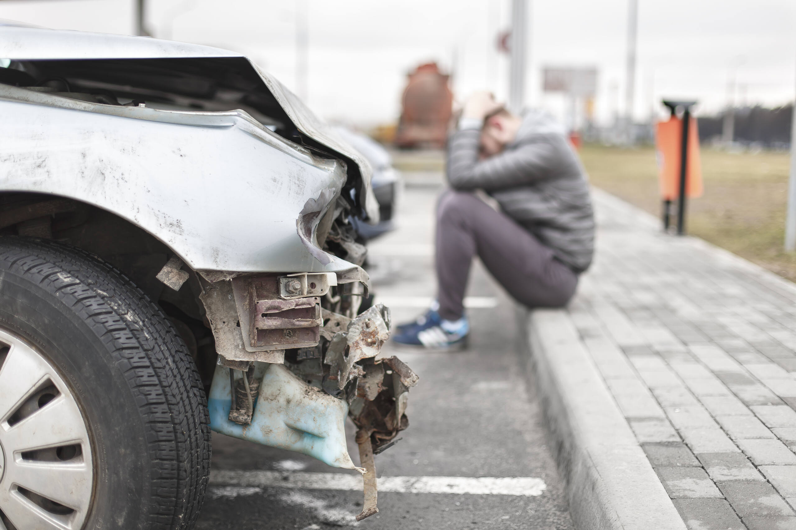 A stressed man sitting on a curb near a crushed bumper questions who is liable in an OKC car accident, the car owner or driver.