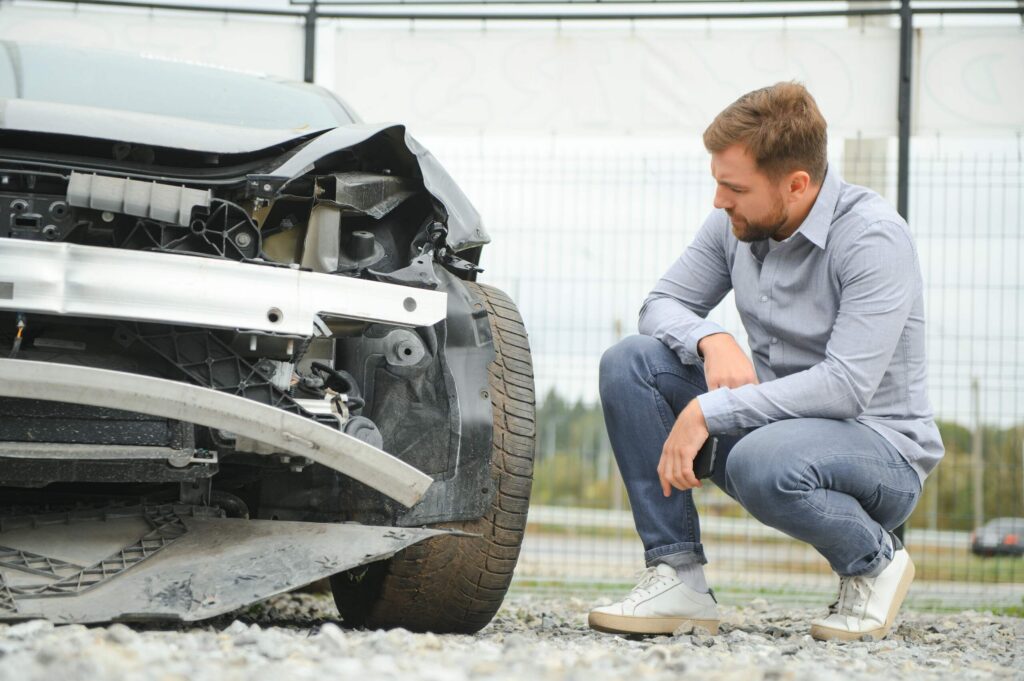A man crouching down to inspect front end damage on his car highlights why get a medical exam after an OKC crash.